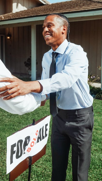 real estate agent standing next to a "For Sale" sign and shaking someone's hand; a "Sold" sticker is affixed to the sign